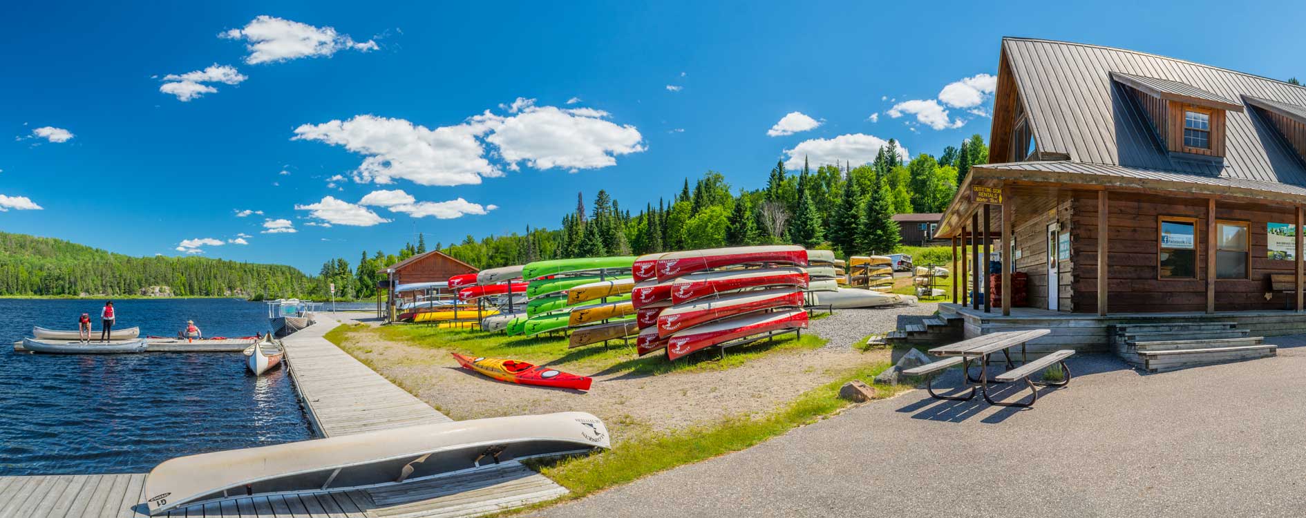 Lake Opeongo Canoe store, a log cabin building on a bright sunny day in summer. There are many colorful canoes set up outside and there are 3 canoers in the water.