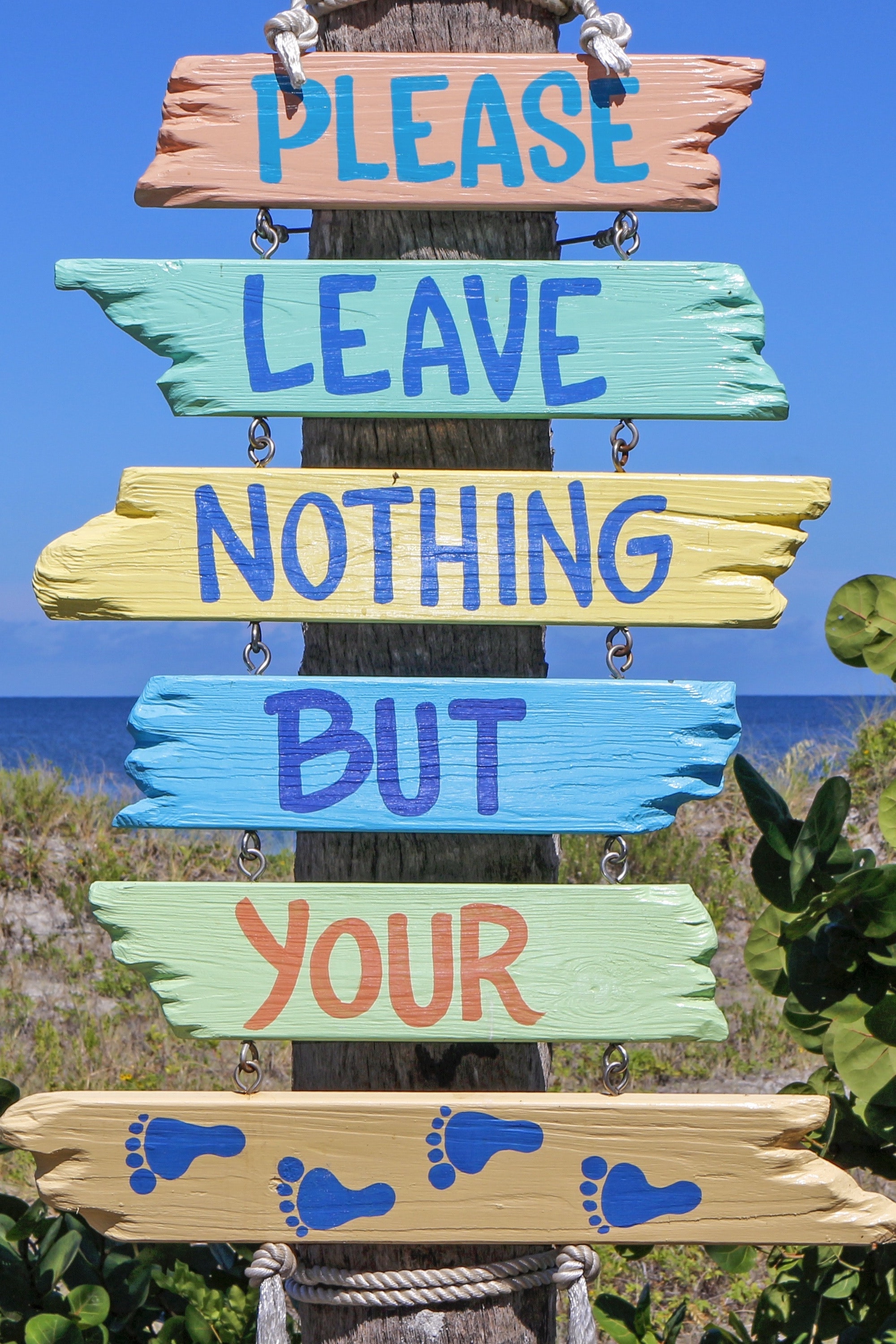 a sign on a beach made of pastel color painted wood planks that says 'please leave nothing but your' and the last plank has painted footprints